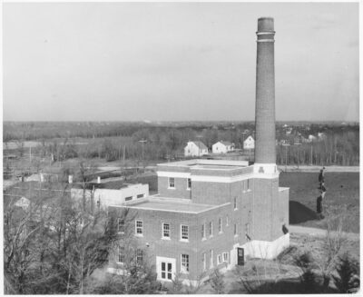 The exterior view of a building with a smoke stack. Houses can be seen in the distance.