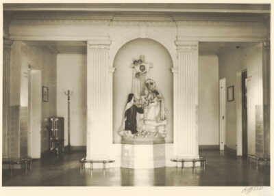 A view of a foyer with ornate columns and sculptures of angels, a nun, and a cross between the columns. Doorways can be seen on either side of the hallway.