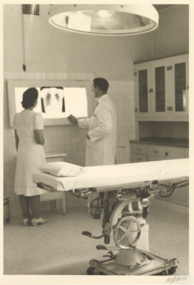A man and a woman look at a chest x-ray on a wall-mounted light box. An operating table sits in the foreground with a large lamp above.