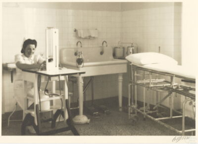 A nurse sits at a table with a tall machine. In the room next to her is a double sink and a gurney.