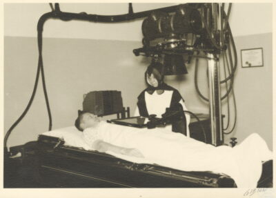 A man lies on a gurney as a nun moves an x-ray plate over his chest. A large machine is suspended above the man.
