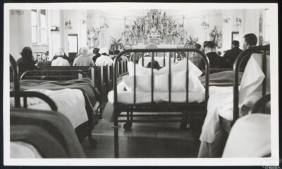 A view from the back of a chapel. People sit in the pews, and hospital beds are gathered behind the pews and down the centre aisle.