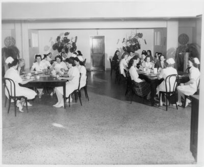 Groups of nurses sit at tables with coffee cups. Large plants stand in the background.