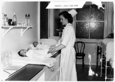 A nurse tends to two infants on a counter. Empty bottles sit on a shelf above the infants and on the counter beside them. A label on the photo reads: "Nursery - Daily Care Room"
