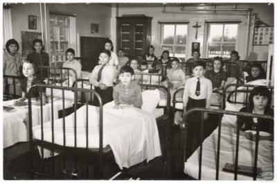 A room filled with children in hospital beds. Other children stand between the beds. A nun can be seen in the background and a cross hangs on the wall.