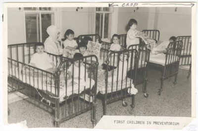 Three cribs and two beds in a line against a wall. Young children fill the beds, and a nun with a white cap sits with the children. A label on the photo reads: "First Children in Preventorium."
