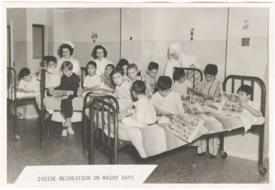 A group of children sit on hospital beds reading newspaper comics. Two nurses and a nun are with the children. A label on the photo reads: "Inside recreation on rainy days."
