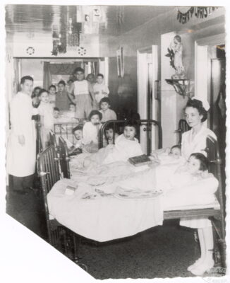 Hospital beds packed tightly in a room with some children lying in the beds and others standing on the beds. A nurse and a doctor stand with the children. Christmas decorations hang on the wall.