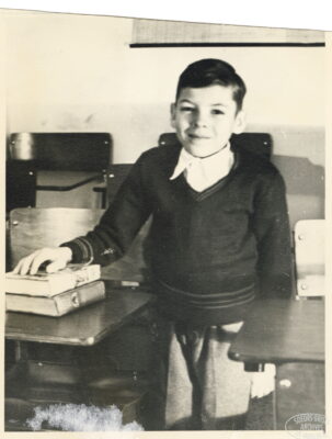 A boy smiles with one hand resting on two books on a desk.