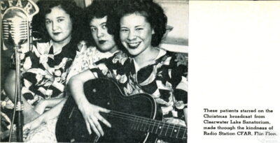 Three women sitting close together behind a microphone that reads "CFAR." The woman on the right holds a guitar. Photo caption reads: "These patients starred on the Christmas broadcast from Clearwater Lake Sanatorium made through the kindness of Radio Station CFAR. Flin Flon."