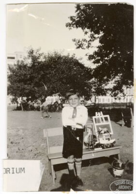 A young boy stands outside and crosses his arms by a bench. A fragment of a label on the photo reads: "orium"