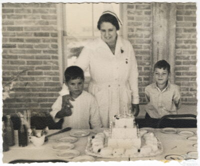 A nurse and two boys stand behind a table with cake. The nurse rests her arm on one boy's shoulder.