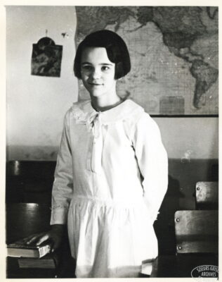 A girl stands next to a school desk and rests her hand on two books. A map hangs on the wall behind her.