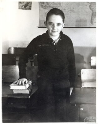 A boy stands next to a school desk and rests his hand on two books. A map hangs on the wall behind him.