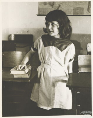 A girl stands next to a school desk, looking to the side, as she rests her hand on two books. A map hangs on the wall behind her.