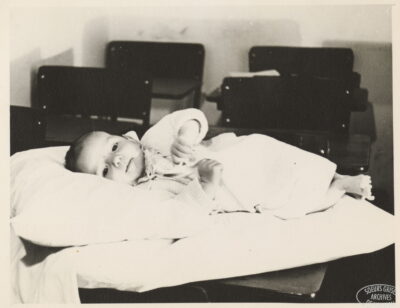 A baby lies on its side on a pillow. Classroom desks can be seen behind the baby.