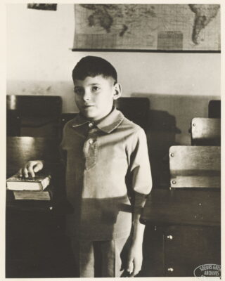 A boy stands next to a school desk and rests his hand on two books. A map hangs on the wall behind him.
