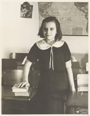 A girl stands next to a school desk and rests her hand on two books. She wears a black top with a white collar and two strings hanging down the front. A map hangs on the wall behind her.
