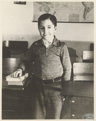 A boy smiles with one hand resting on two books on a desk. A map hangs on the wall behind him.