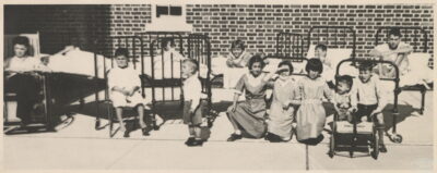 A group of children outside near a brick building. Some children are in hospital beds, some are in wheelchairs.