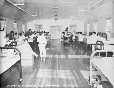 Children in hospital beds lined up against the left and right walls. A nurse and two men can be seen amongst the children. An annotation on the photo reads: "RE 5477-2 Clearwater Lake Sanatorium 21 Sept 50."