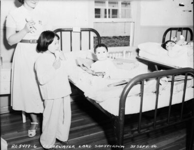 A woman and a child stand next to two young children in hospital beds. The woman and two children hold ice cream cones, and one child holds a toy. An annotation on the photo reads: "RE-5477-6 Clearwater Lake Sanitorium 21 Sept 50"