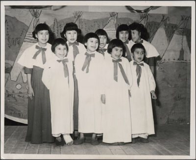 A group of nine children grouped together in front of a painted background depicting a camp scene with tipis.