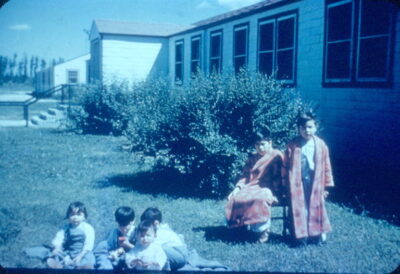 A group of toddlers sit on a blanket on the grass in front of a building, next to two boys wearing robes.