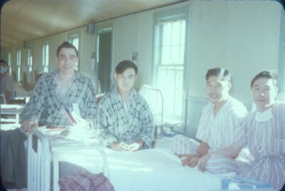 Four men sitting on hospital beds. Two men are wearing plaid pyjamas and two men are wearing striped pyjamas.