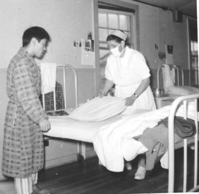 A nurse makes a bed with the help of a man in a plaid robe.