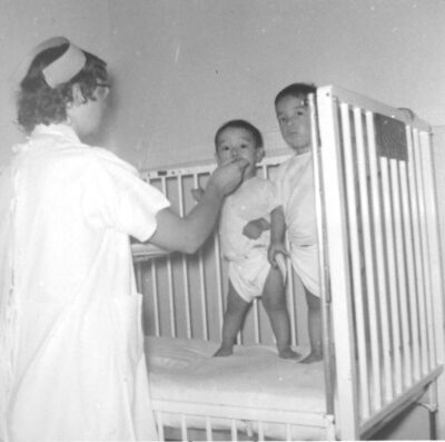 Two toddlers stand in a crib. A nurse feeds one of the the toddlers.