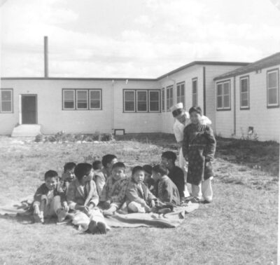 A group of boys sit on the grass in front of a building. A nurse rests her arm on the shoulder of a boy who stands next to the other boys.
