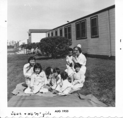 Six children sit on the grass outside of a building. A nurse kneels next to them and holds another child. An annotation on the bottom of the photo reads, "Jean & WD "M" girls"