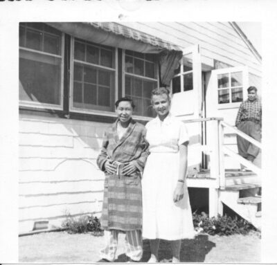 A young man in a plaid robe stands next to a nurse outside the entrance of a building. Another man stands at the doorway.