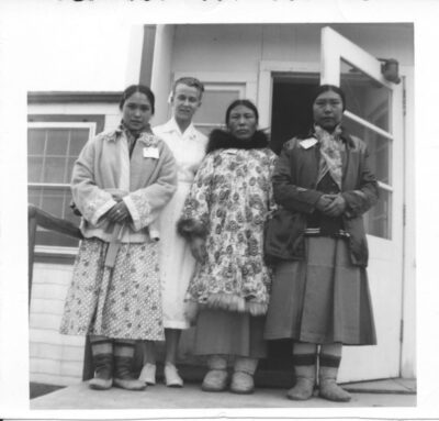 A nurse and three women wearing skirts, coats, and boots stand at the doorway of a building.