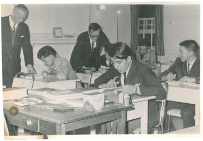 Five men sitting at desks in a classroom with two other men standing beside them