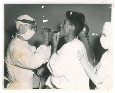Man receiving oral medical care from male doctor with female nurse supporting patient from behind. Stamp in lower right reads "Larry"
