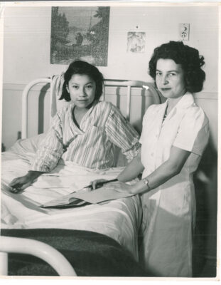 A nurse stands next to a teen girl in bed, holding writing materials