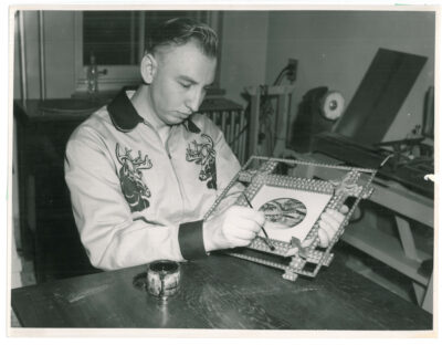 A man painting the frame of a picture. He is wearing a zippered jacked with two elk pictured on the chest.