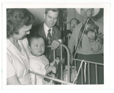 Toddler standing in a crib holding a small guitar surrounded by nurse, a man playing a violin, and two other men. One man appears to be wearing a fraternal order hat that reads "The Pas." Another child lays in a crib in the background