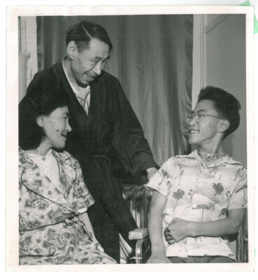 A woman and young man sit in chairs and smile at each other. A man stands behind them and smiles at the young man.