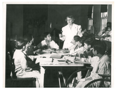 Boy patients around a table writing with nurse teacher. One boy sits in a wheelchair.