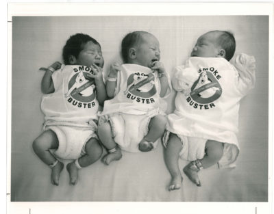 Three babies with medical bracelets on wrists and ankles, wearing "Smoke Buster" shirts and cloth diapers. The shirts are a play on the ghost busters logo with "The Manitoba Lung Association" on the logo