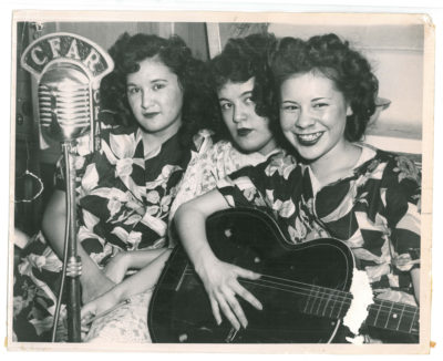Three women sitting close together behind a microphone that reads "CFAR." The woman on the right holds a guitar.