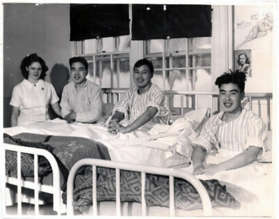 Three smiling men sitting in bed, with a nurse standing next to them.