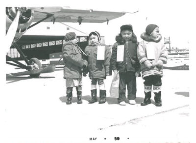 Four young children in front of airplane. They have large tags pinned to their coats.