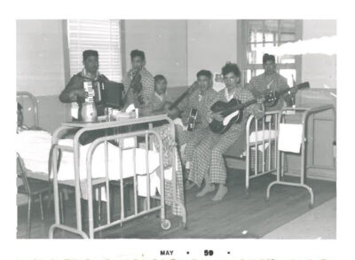 Six men in plaid pyjamas sit on a hospital bed and play musical instruments. One man plays an accordion, another plays a violin, and the remaining men play guitars.