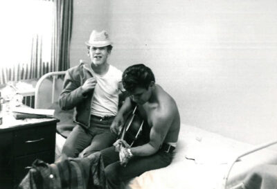 Two young men sitting on hospital bed. One man is wearing a fedora hat and is putting on a jacket. The other man is shirtless and is playing a guitar.