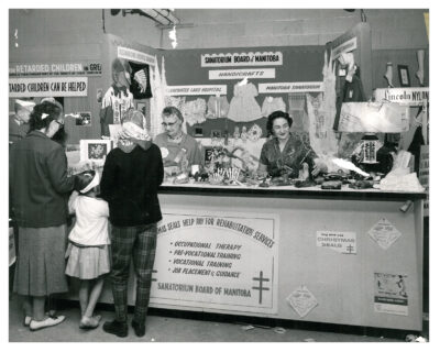Two women working at a booth displaying handicrafts on behalf of the Sanatorium Board of Manitoba. Two women and a child stand in front of the booth.