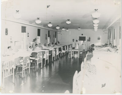 A hospital ward with several men in beds lined up along the walls, and five medical staff standing at the back of the room. Stamp: "Larry"